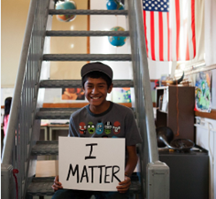 Boy at school sitting on a staircase holding a sign that reads 