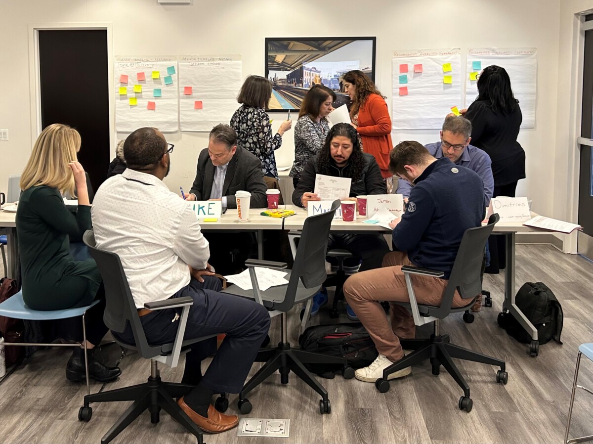Staff working on their SEL activities. Some are circled around a table writing. Others are looking at large sticky pad sheets that have notes and smaller sticky pads attached as a gallery. 