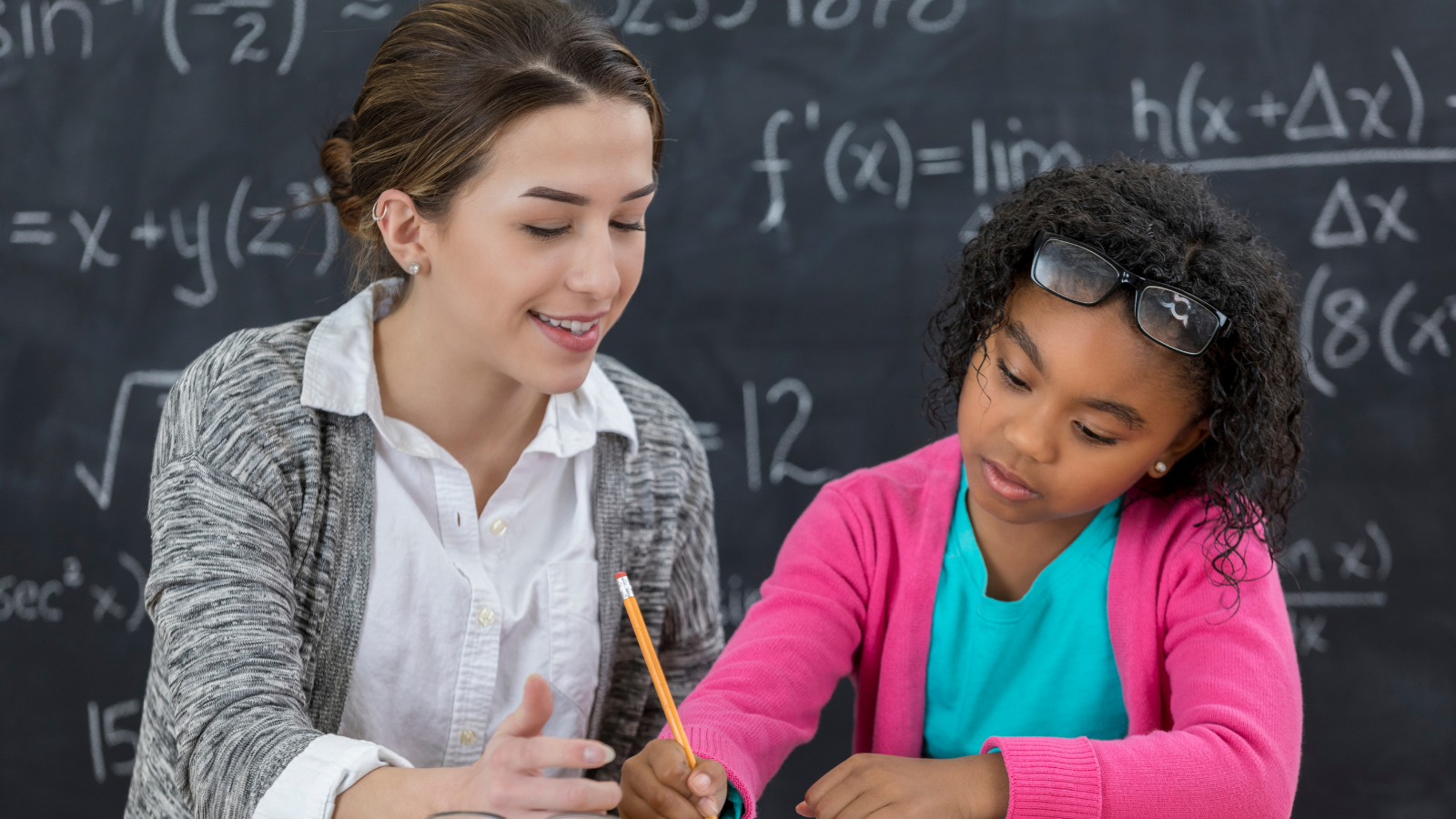 Teacher working with a student. A black board behind them shows math equations.