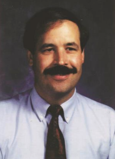 Joseph E. Zins - A light-skinned, male-presenting person with a mustache wearing a tie