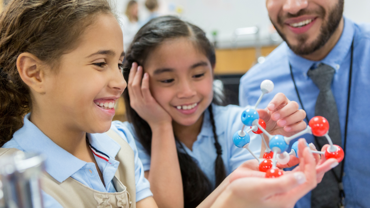 2 female students working with a model of an atom while a male teacher watches