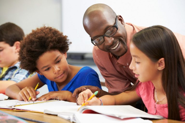A male-presenting person helps several students at a table in a classroom.